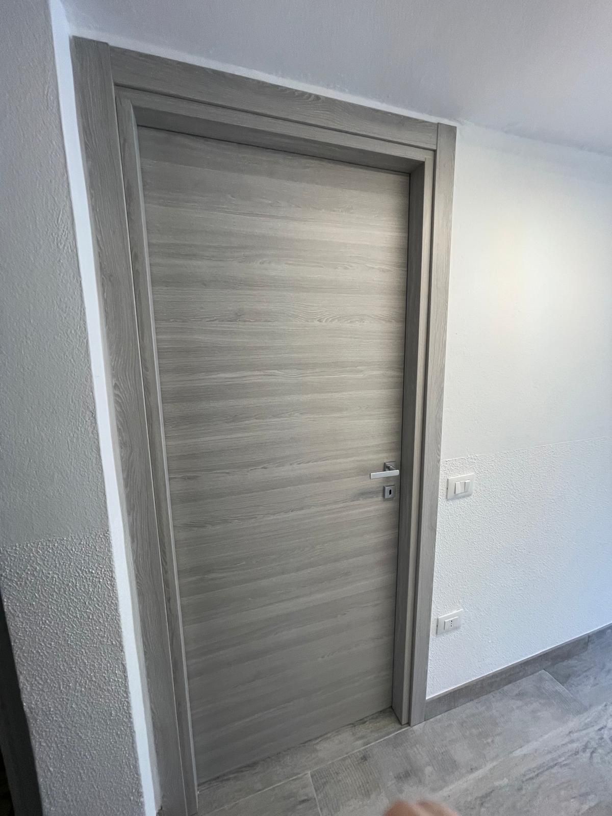 Porta chiusa con finitura effetto legno grigio in un corridoio bianco luminoso con pavimento piastrellato.