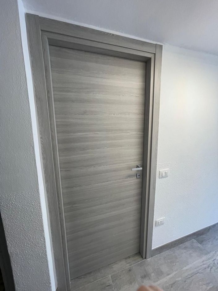 Porta interna chiusa in legno chiaro in un corridoio bianco con pavimento in piastrelle grigie.