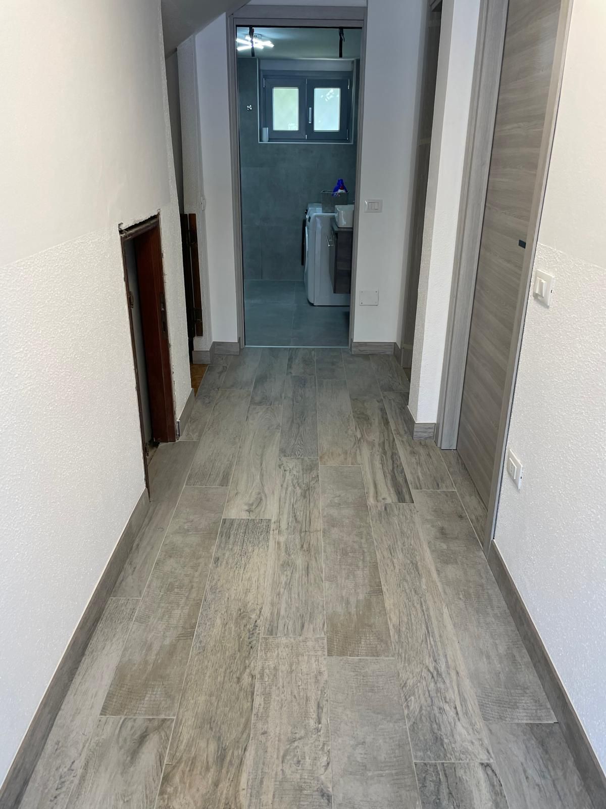 Stretto corridoio con pavimento in legno grigio che conduce a un piccolo bagno o ripostiglio in fondo.