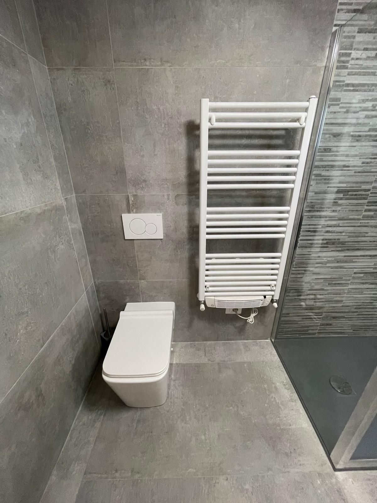 Bagno moderno con piastrelle grigie, WC sospeso, termoarredo bianco e pannello doccia in vetro.