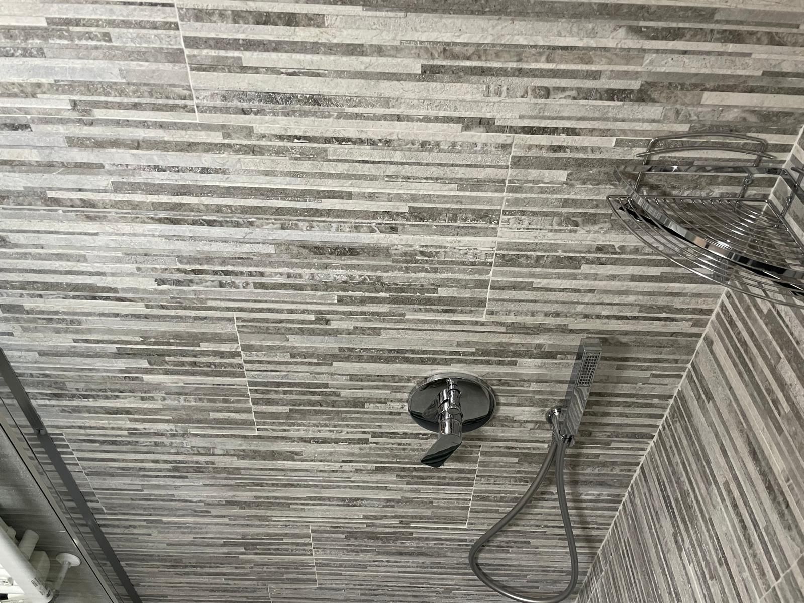 Angolo doccia con pareti rivestite in piastrelle grigie, rubinetteria cromata e doccetta a mano con tubo flessibile.