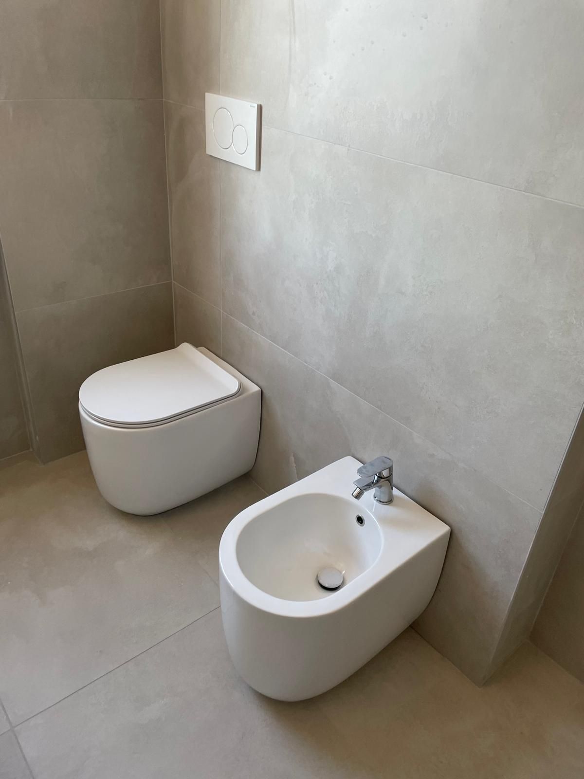 Bagno moderno con WC e bidet, pareti grigie e pavimento piastrellato.