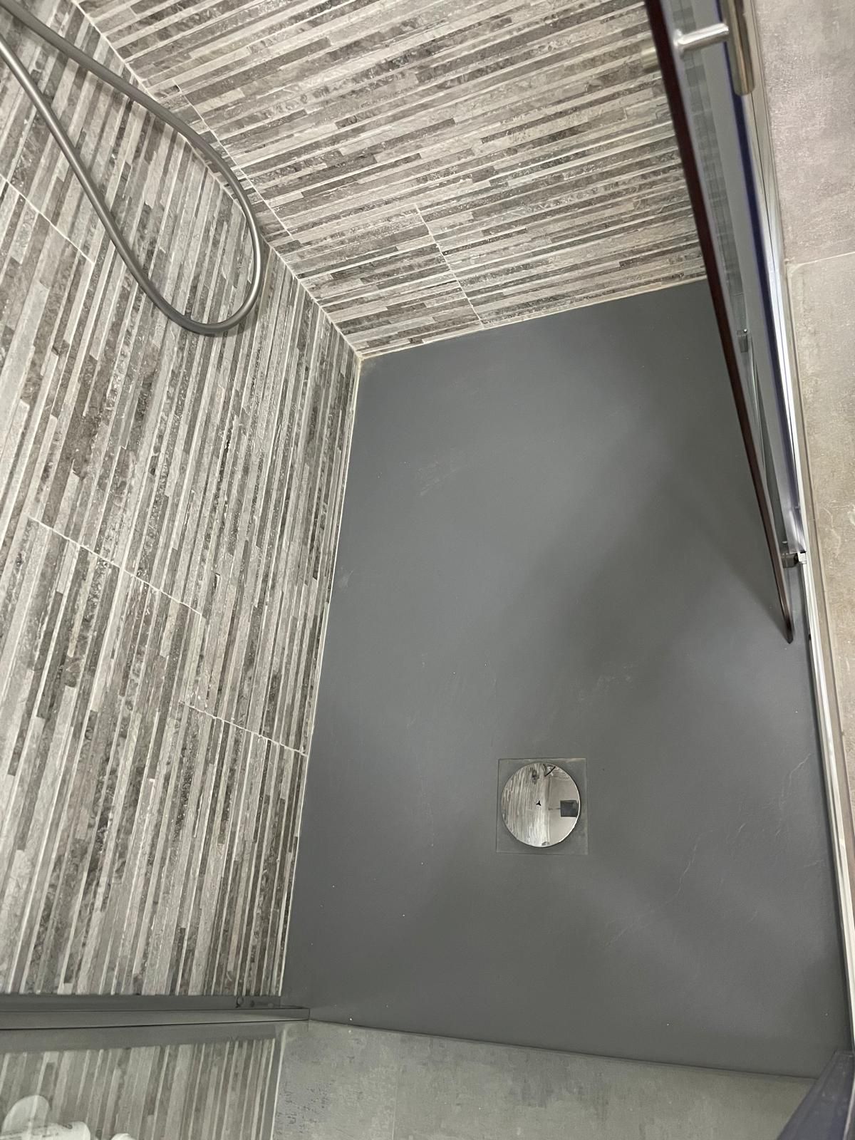 Pavimento della doccia grigio con scarico centrale accanto a piastrelle a parete effetto legno e porta doccia in vetro.