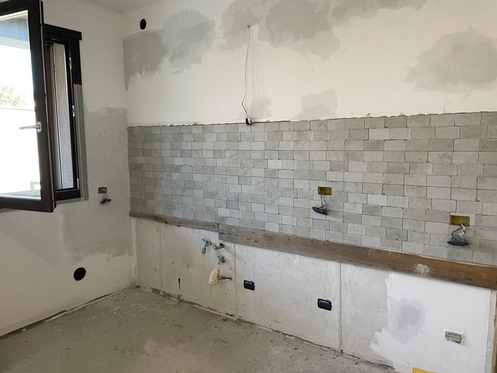 Cucina non rifinita con pareti bianche, paraschizzi piastrellato e apertura per finestra sulla sinistra.