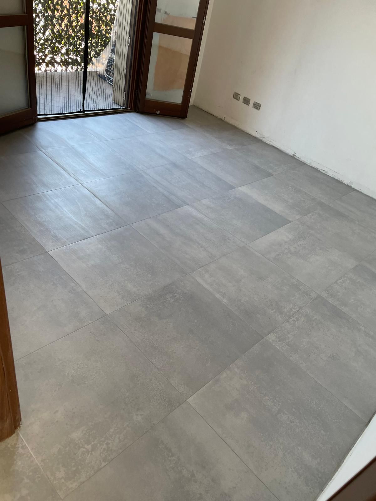 Stanza vuota con pavimento piastrellato grigio chiaro, porte in legno e luce solare proveniente da una porta-finestra aperta sul balcone.