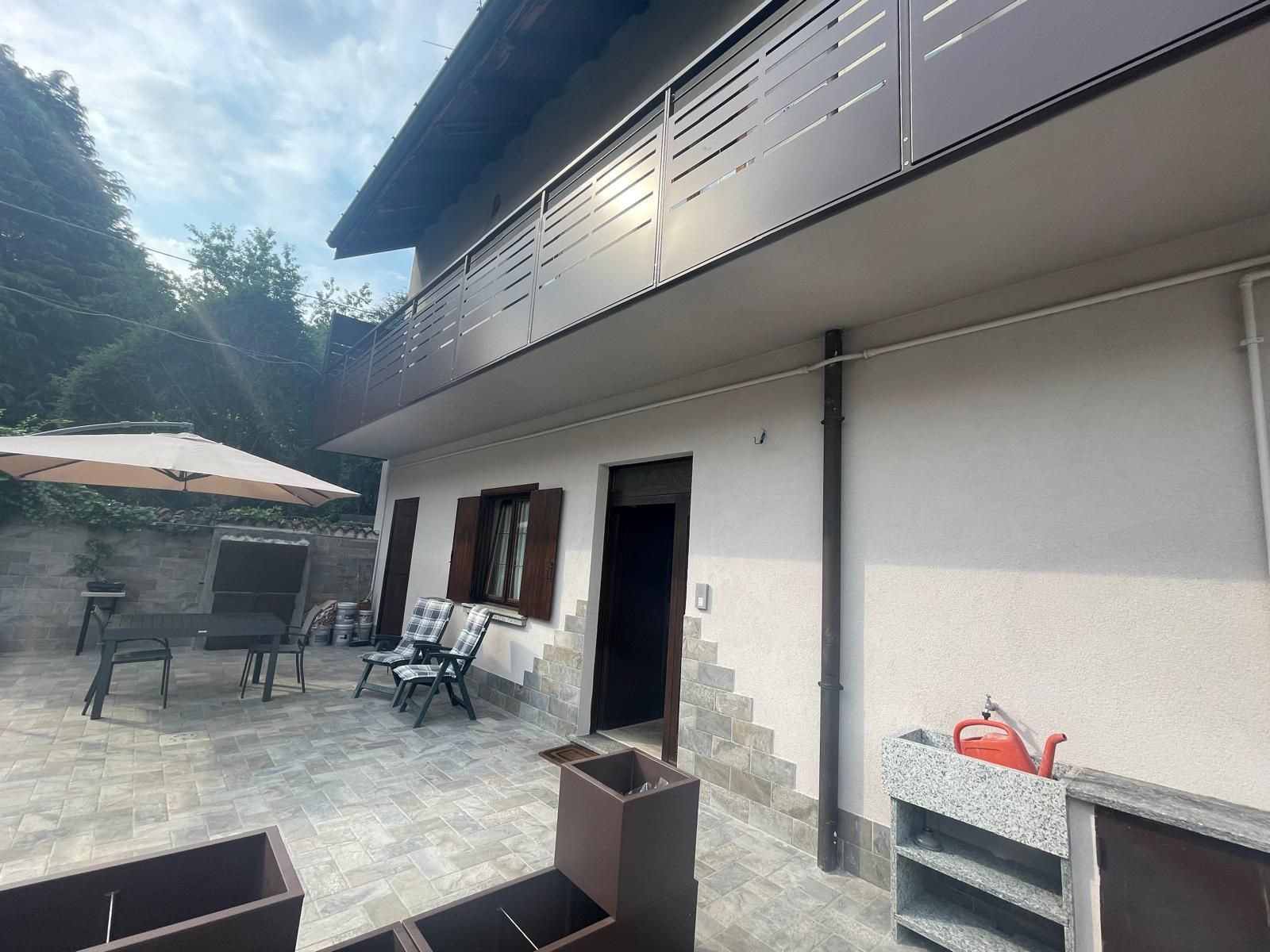 Patio esterno accanto a una casa bianca con tavoli, sedie e una porta aperta sotto un balcone scuro.