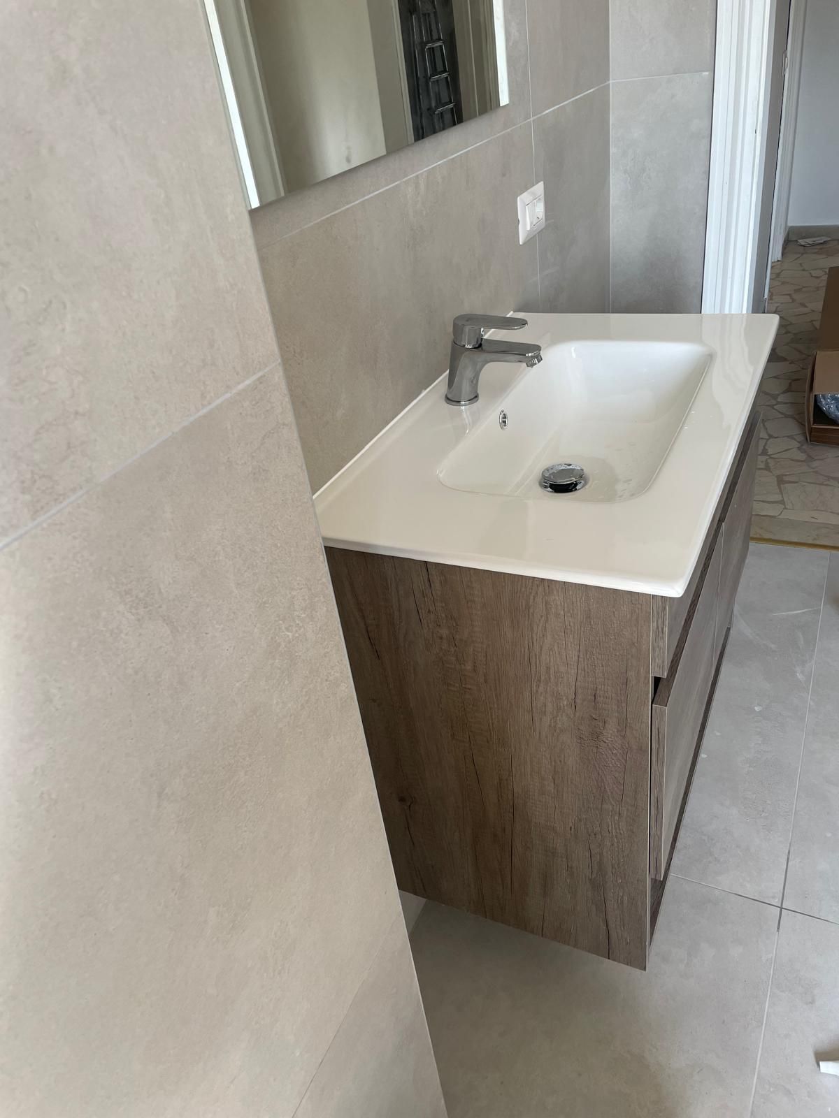 Mobile bagno moderno con lavabo bianco, rubinetto cromato e mobiletto in legno accanto a uno specchio e pavimento piastrellato.