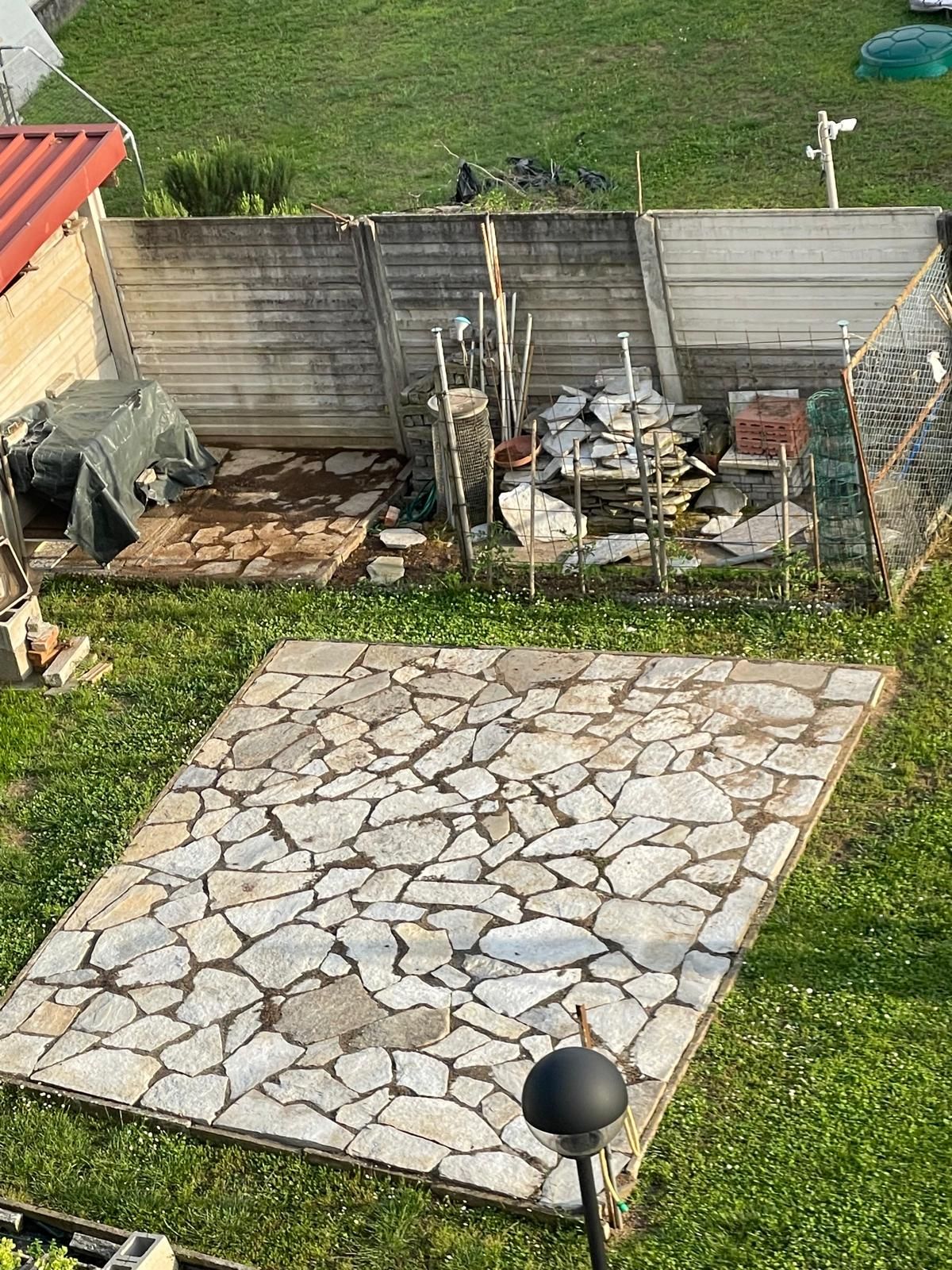 Patio in pietra in un cortile recintato con piante in vaso e attrezzi da giardino lungo il muro.