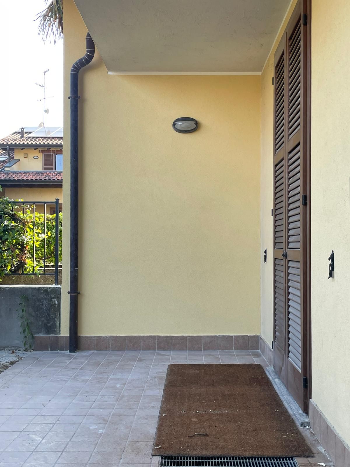 Veranda coperta con parete beige, pavimento piastrellato, applique e zerbino accanto alle porte a persiana.