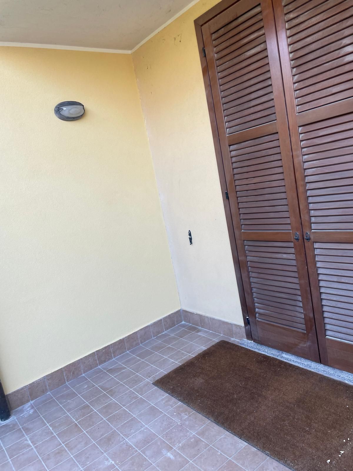 Piccolo portico piastrellato con pareti beige, zerbino marrone e porta a persiana in legno.