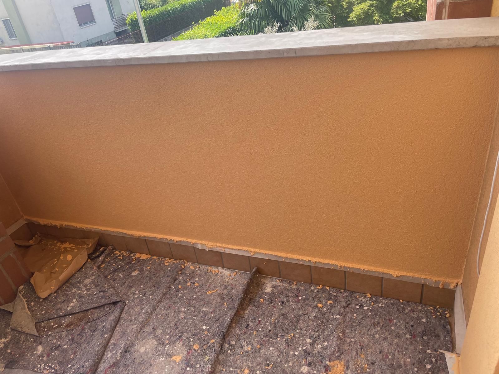 Angolo vuoto del balcone con parete arancione, pavimento piastrellato e un piccolo cumulo di detriti vicino al lato sinistro.