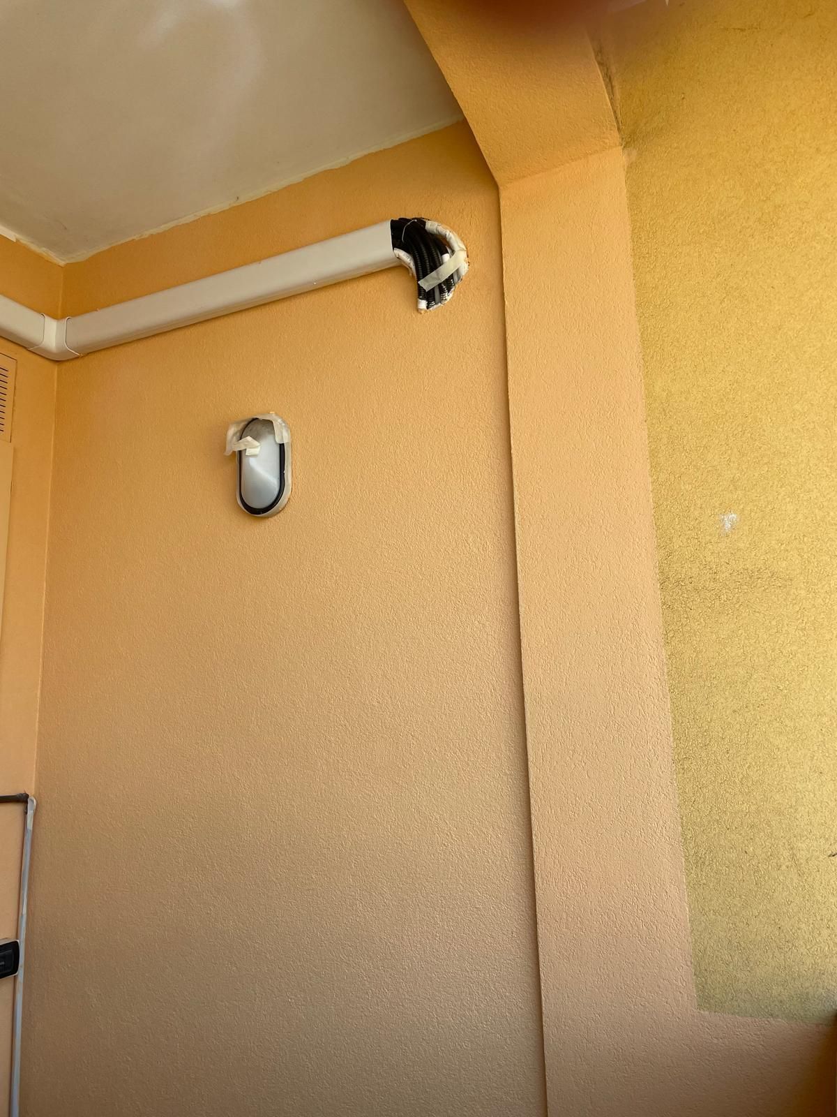 Angolo di parete esterna beige con lampada a sospensione e piccola apertura nera vicino al soffitto.