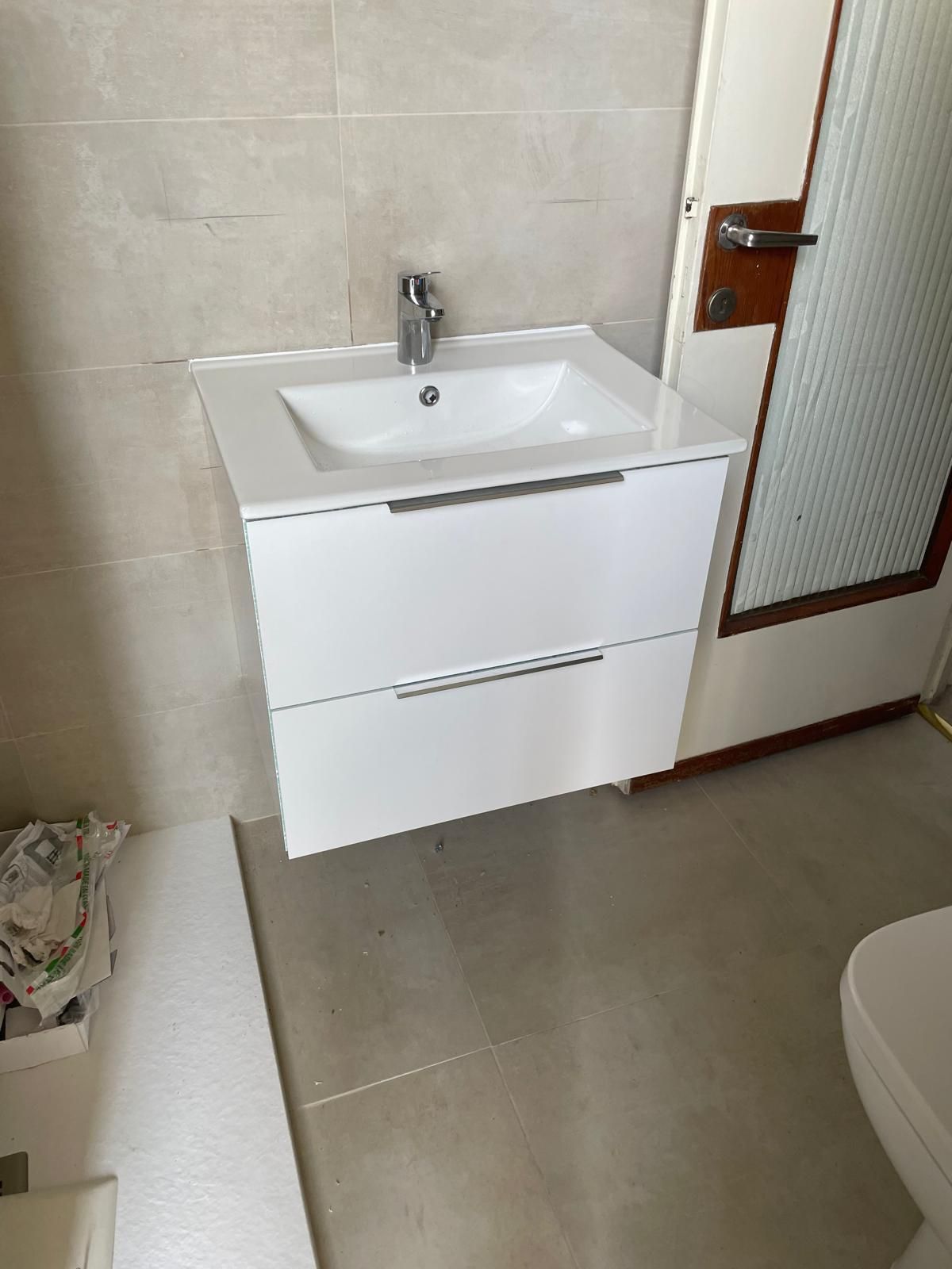 Mobiletto da bagno di piccole dimensioni con lavabo e specchio bianchi, pareti rivestite con piastrelle beige e anta in vetro satinato.