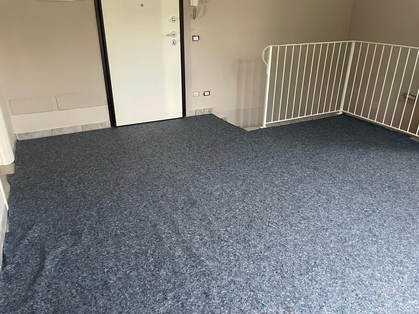 Stanza vuota con moquette blu, pareti bianche, una porta e una ringhiera bianca sul lato destro.