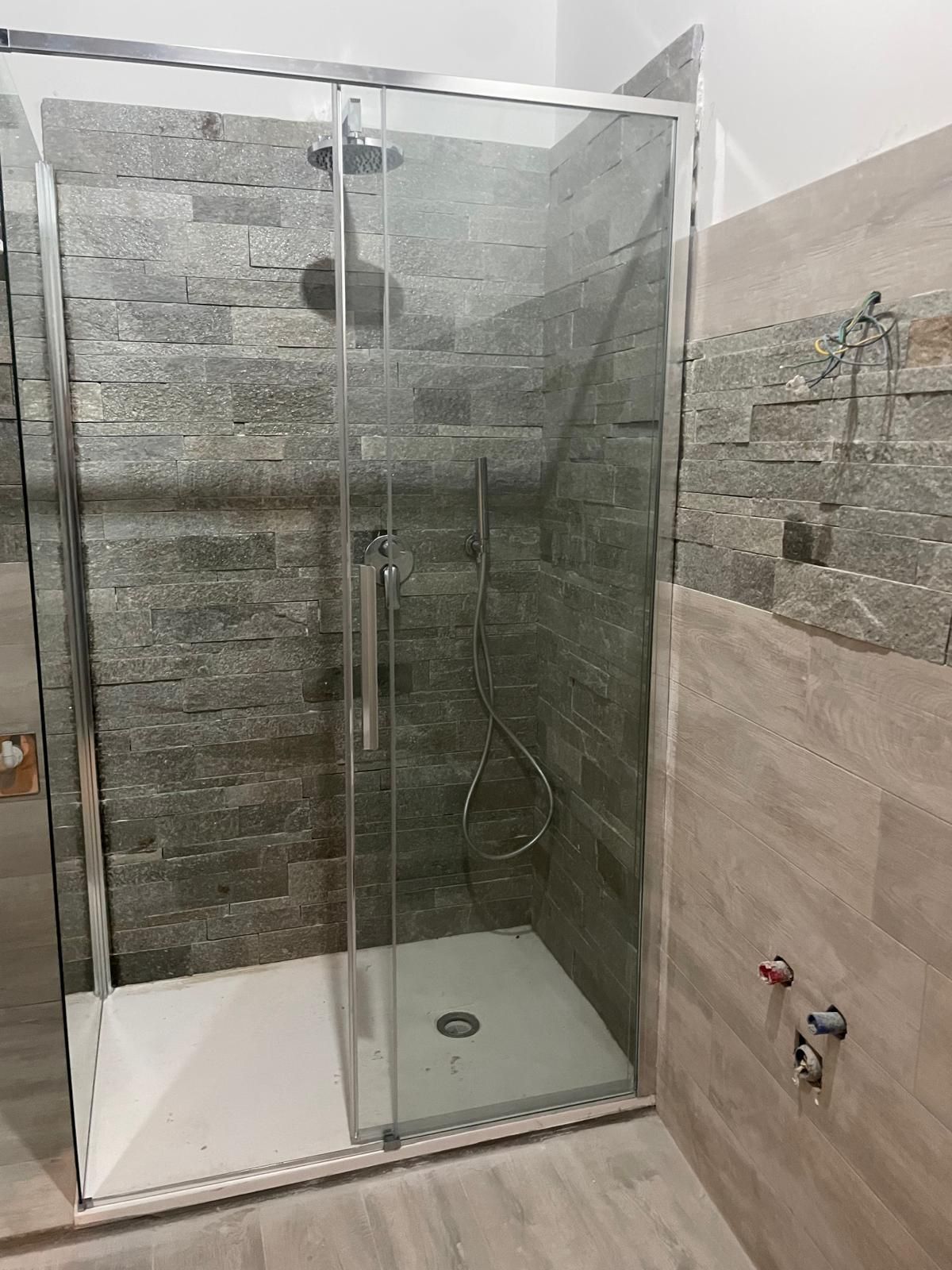 Box doccia con pareti in vetro, rivestimento in piastrelle di pietra, piatto doccia bianco e soffione a parete.