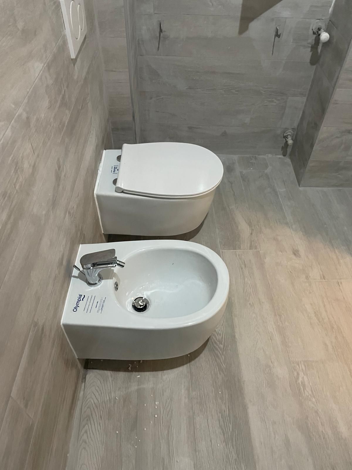 Piccolo bagno con bidet e WC bianchi su pavimento piastrellato grigio.