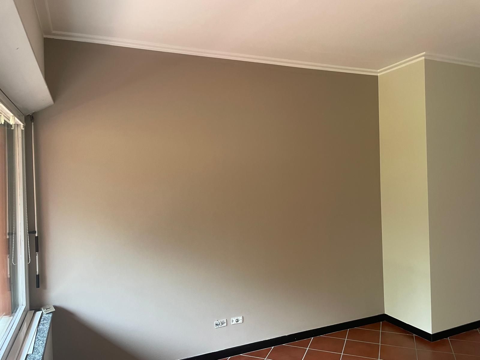 Stanza vuota color beige con grande finestra, parete grigia, battiscopa scuri e pavimento piastrellato.