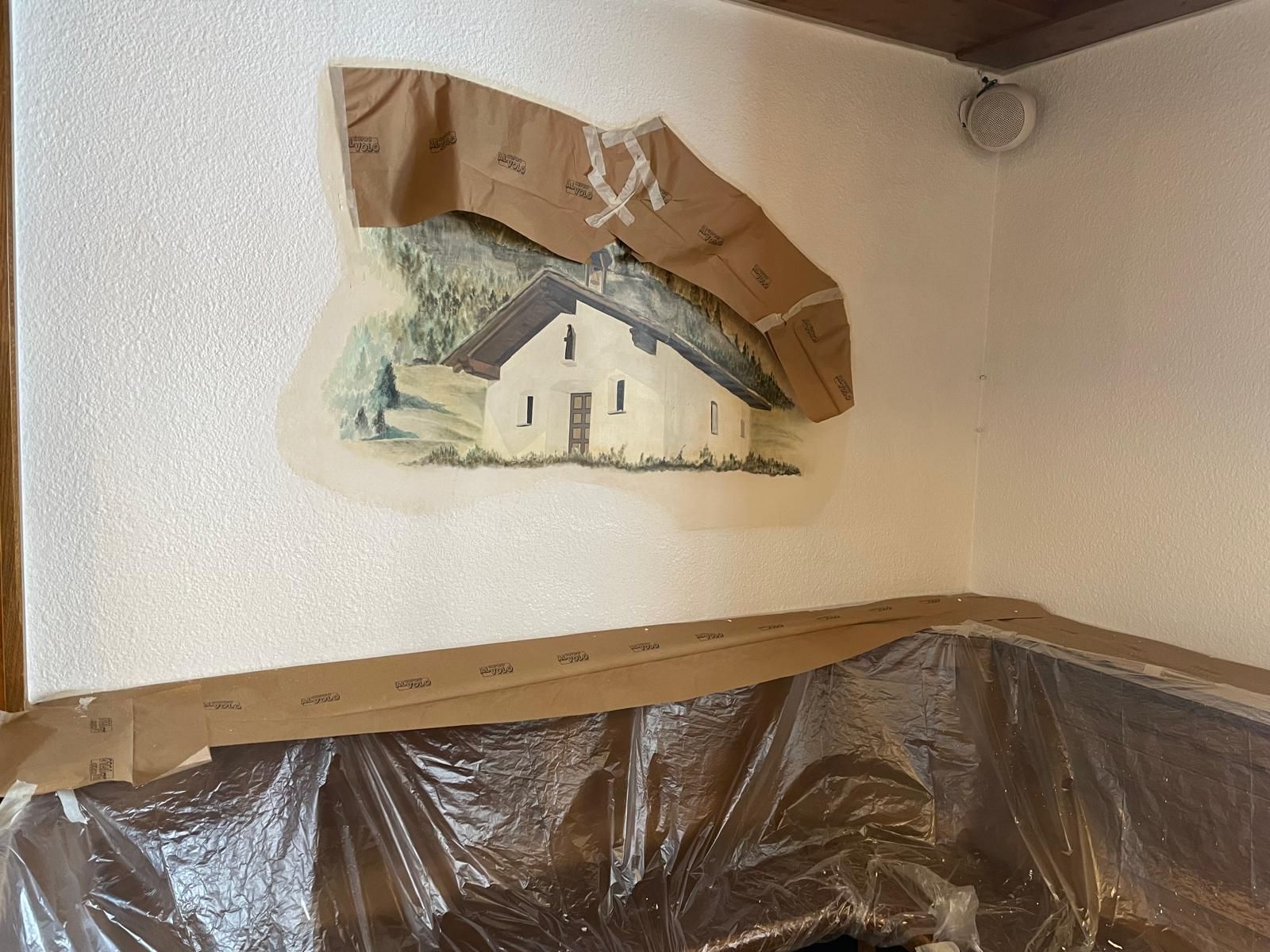 Angolo di una stanza con teli di plastica protettivi e un murale che viene riportato alla luce durante i lavori di ristrutturazione.