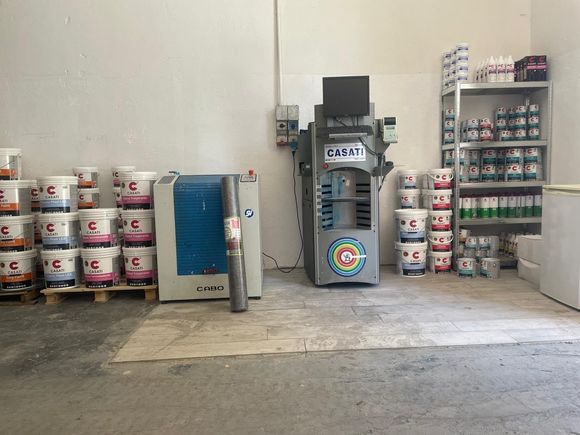 Corridoio del magazzino con secchi di vernice impilati, una macchina per miscelare e scaffalature metalliche.