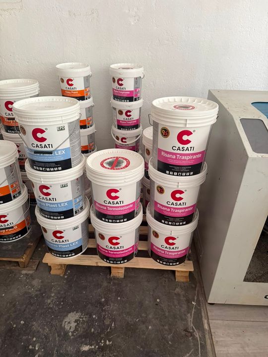 In un'officina, accanto a un macchinario, sono impilati dei secchi di vernice bianca con etichette rosa e blu su un pallet.