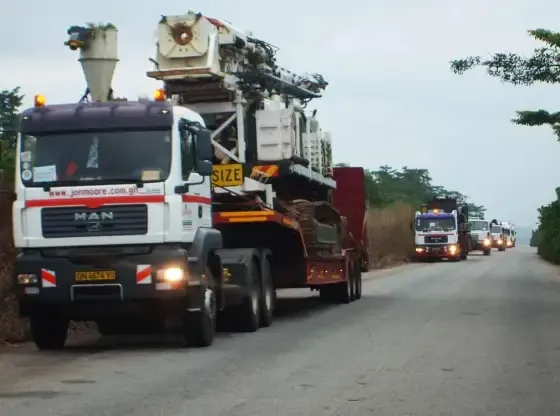 Line of heavy trucks carrying large machinery drives along a rural road at dusk