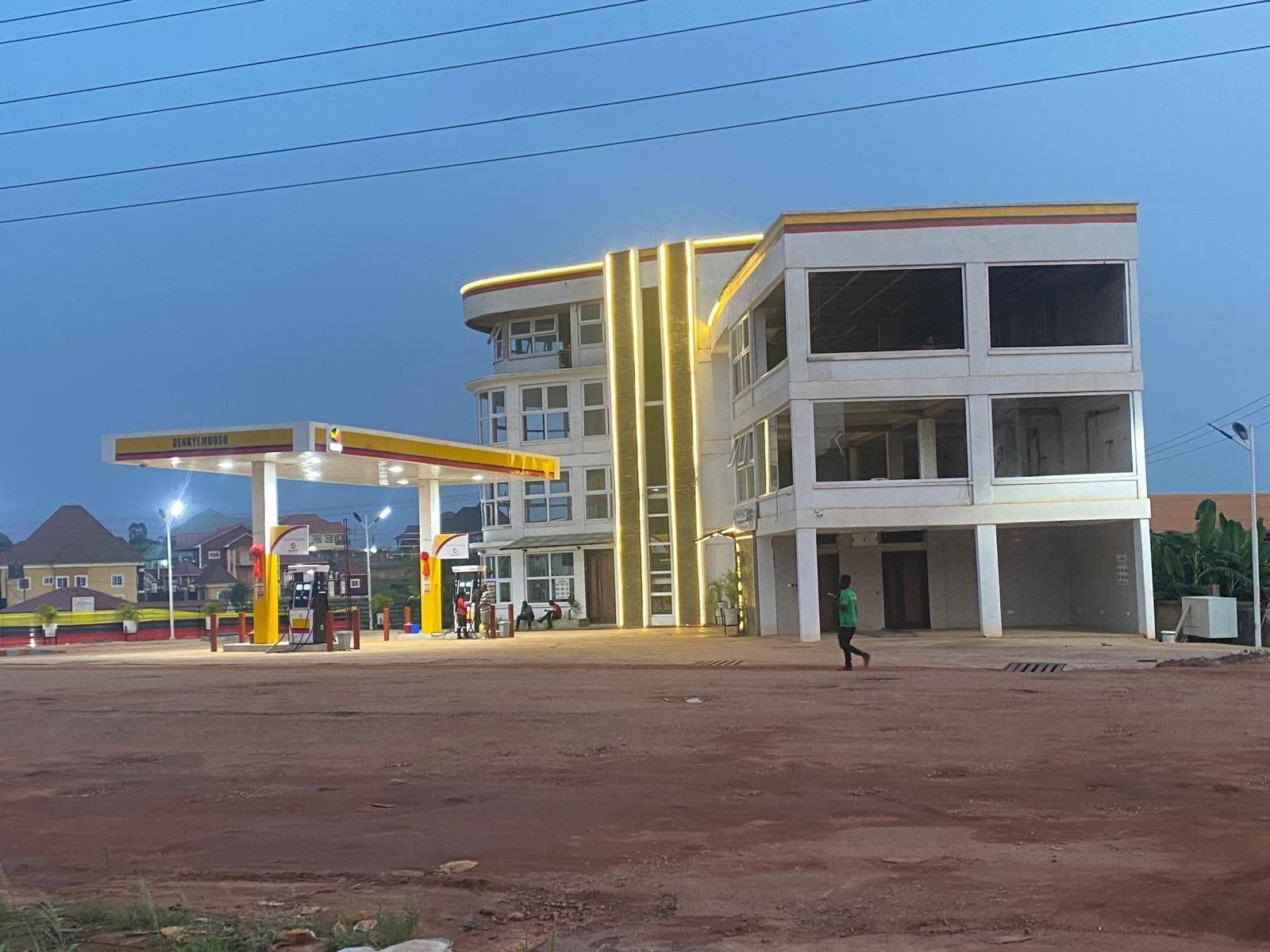 Modern gas station at dusk with a lit canopy and a white multi-story building beside a dirt lot.