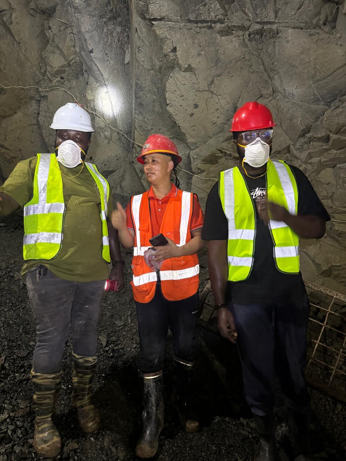 Three workers in hard hats and safety vests standing in a rocky tunnel.