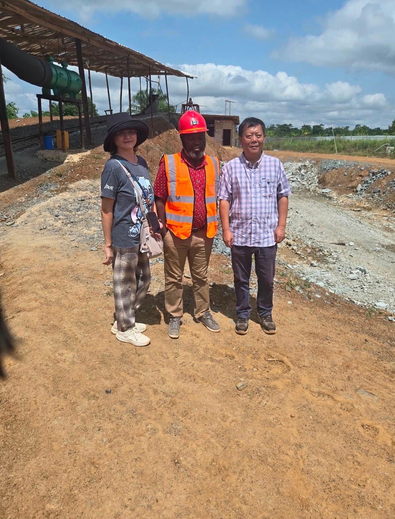 Three people standing on a dirt road at a construction site under a partly cloudy sky