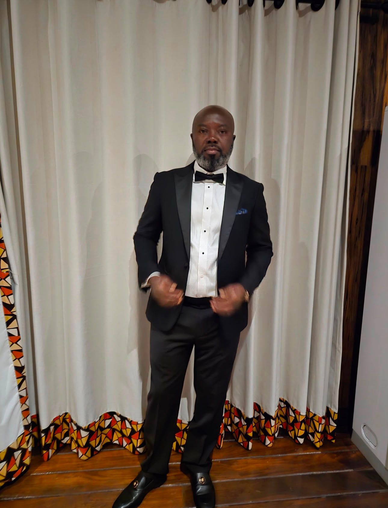 Man in a black tuxedo posing in front of white curtains and patterned trim.