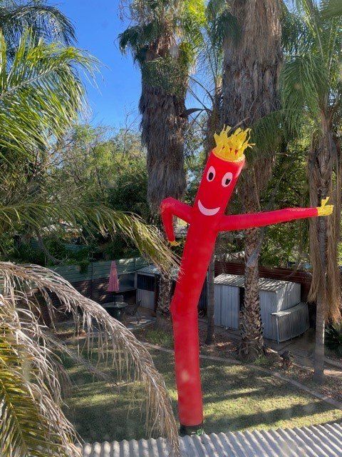 A red inflatable tube man with a smiling face and crown dances in a yard with palm trees.