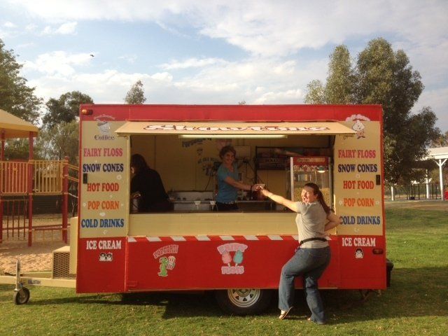 A woman gestures excitedly near a red food trailer selling ice cream and snacks in a park. Two people work inside.