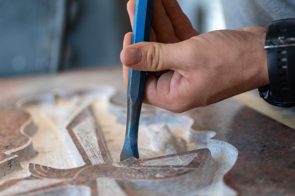 A hand chiseling details into a stone surface with a blue-handled tool. The carving features light-colored lines.