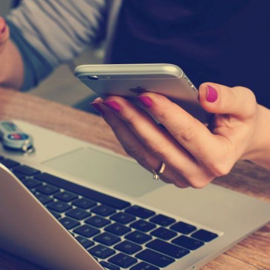 Person using a smartphone, with the phone in hand over a laptop. The person's nails are painted pink.