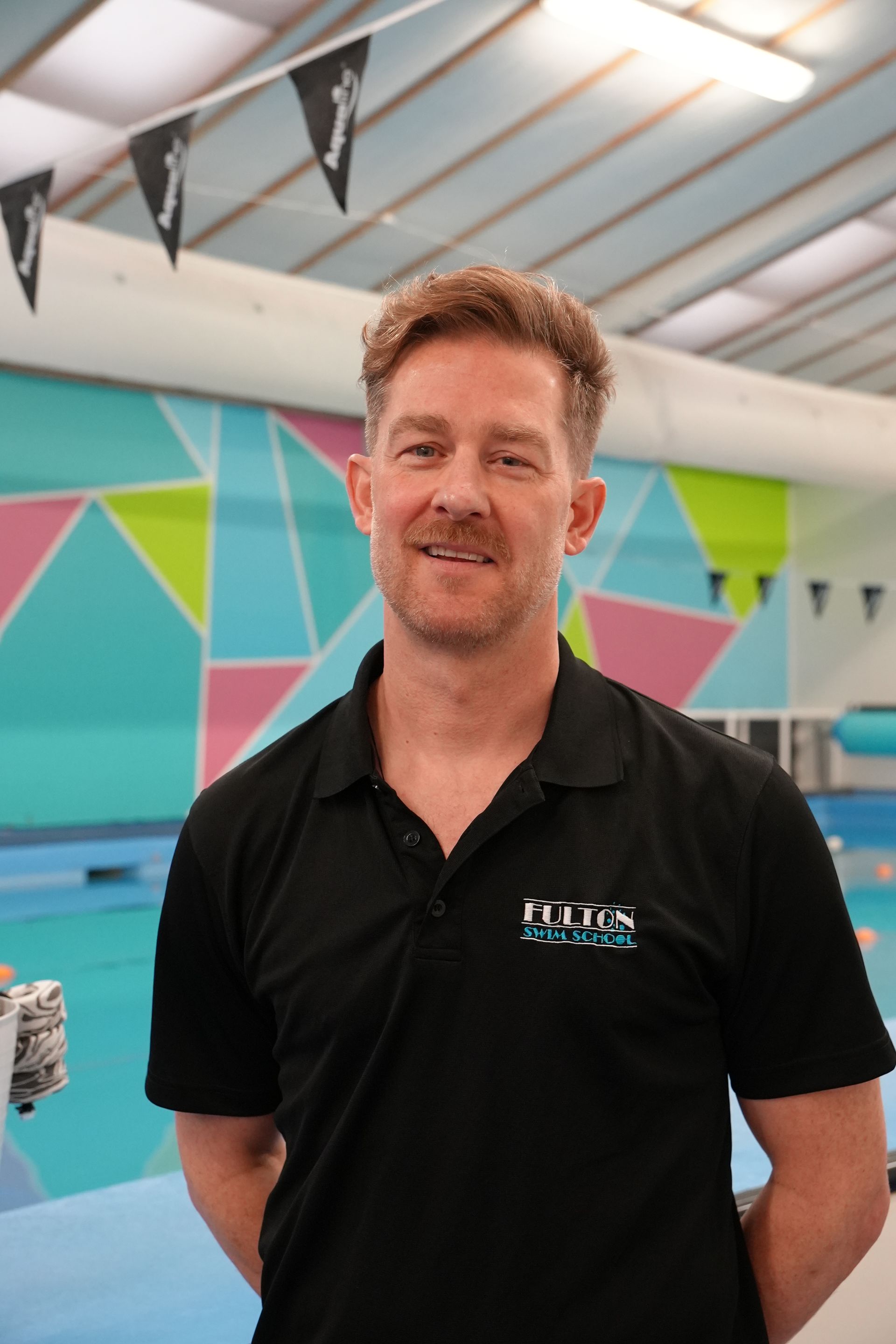 A swim teacher in a black shirt is standing in front of a swimming pool