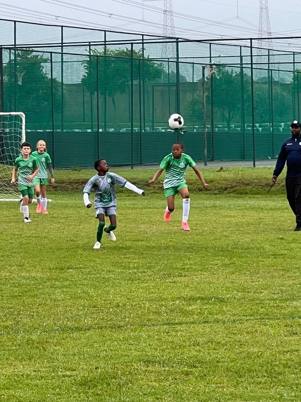 Children in soccer uniforms play on a green field, with one child jumping to head a soccer ball toward a goal.
