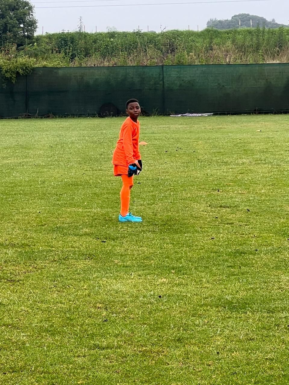 A person in an orange soccer uniform stands on a green grass field near a dark fence.