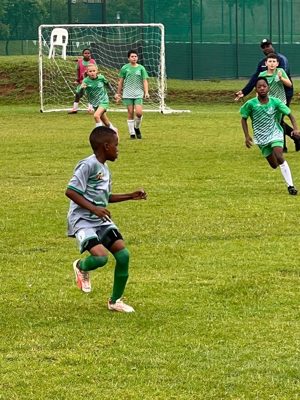 Youth soccer players in green and white uniforms play on a field near a goal net.