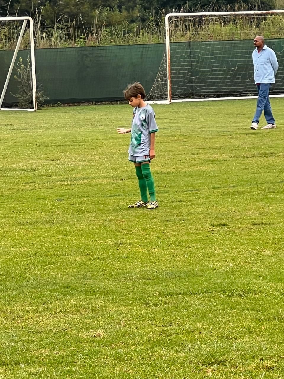 A child in a grey soccer uniform and green socks stands on a grassy field near a goal, as an adult walks in the distance.