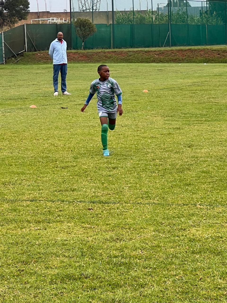 A child in a patterned gray soccer kit runs across a grassy field while a coach observes in the background.