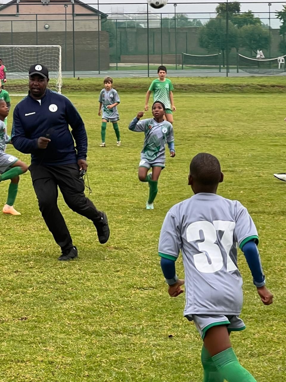 A coach in a dark blue tracksuit watches youth soccer players in grey and green uniforms practicing on a grassy field.