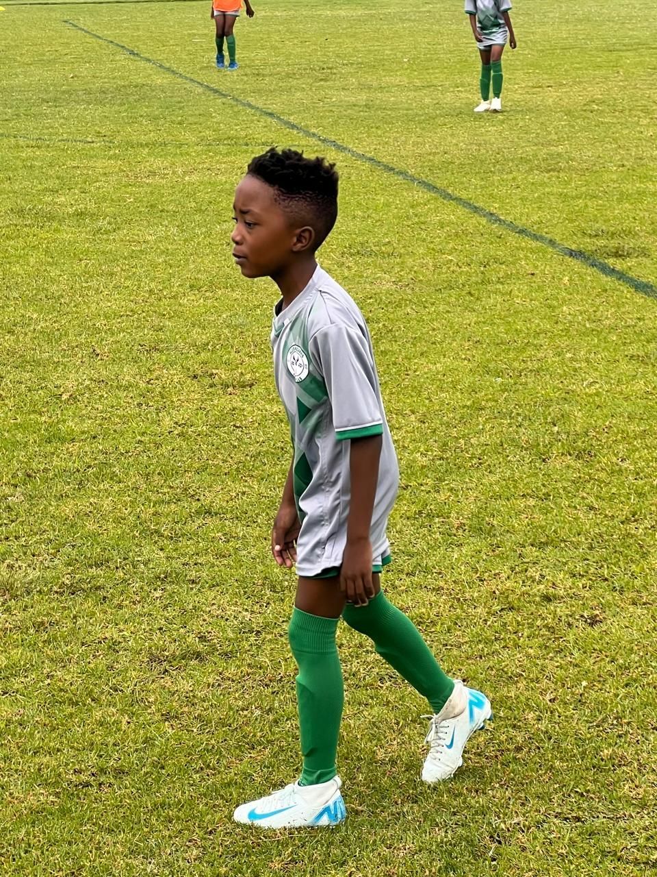 A person wearing a grey and white soccer jersey and green socks walks across a grassy field.