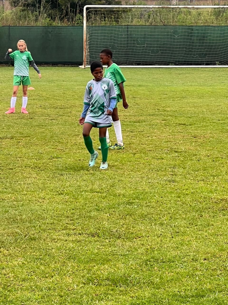 Three children in athletic uniforms play soccer on a green grass field near a goal post.