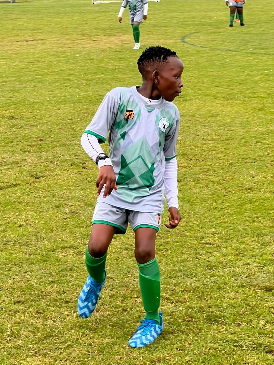 A soccer player in a grey and green patterned uniform and blue cleats stands on a grass field during a match.