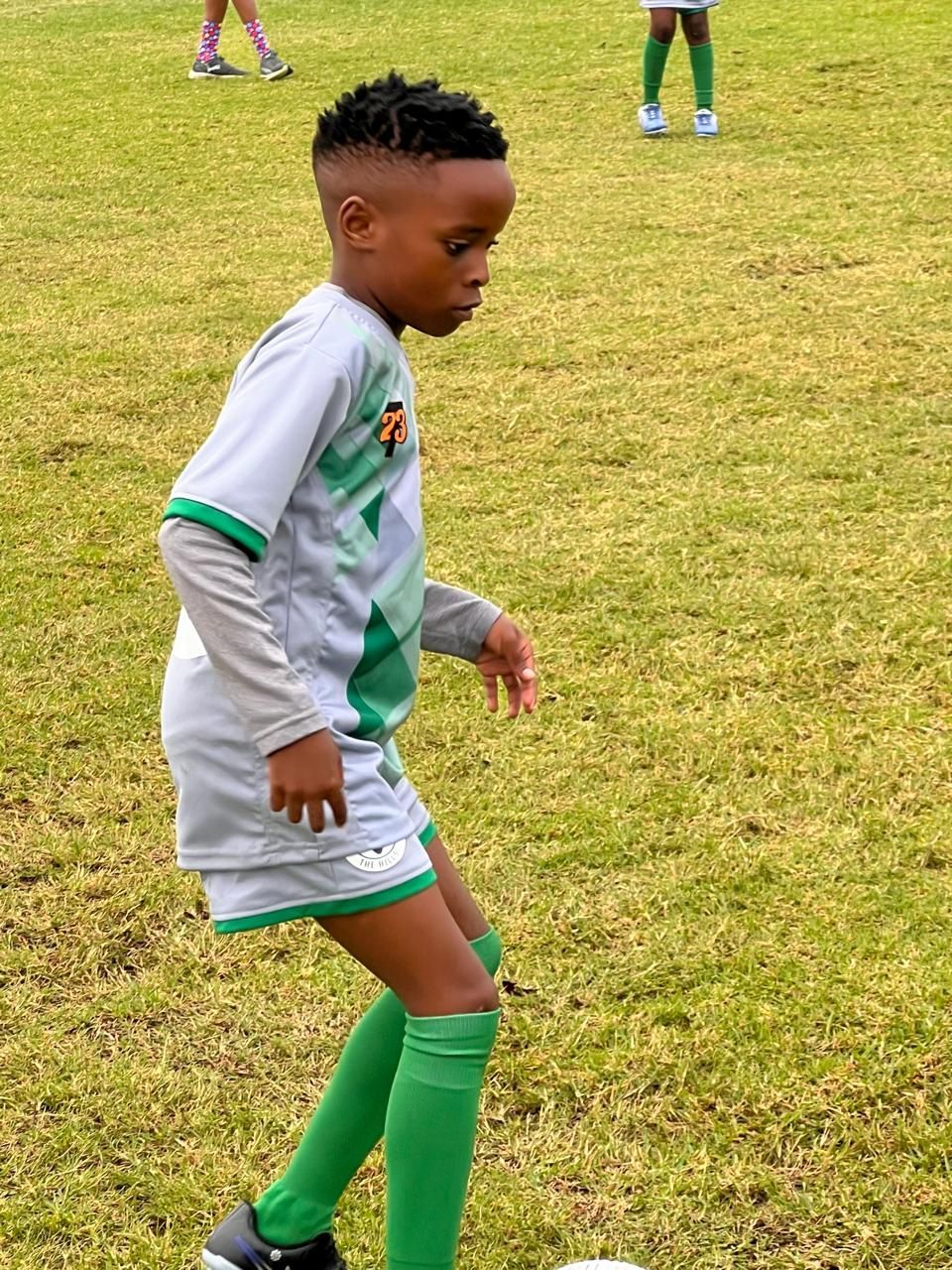 A child in a grey and green soccer uniform stands on a grass field, focused on a ball at their feet.