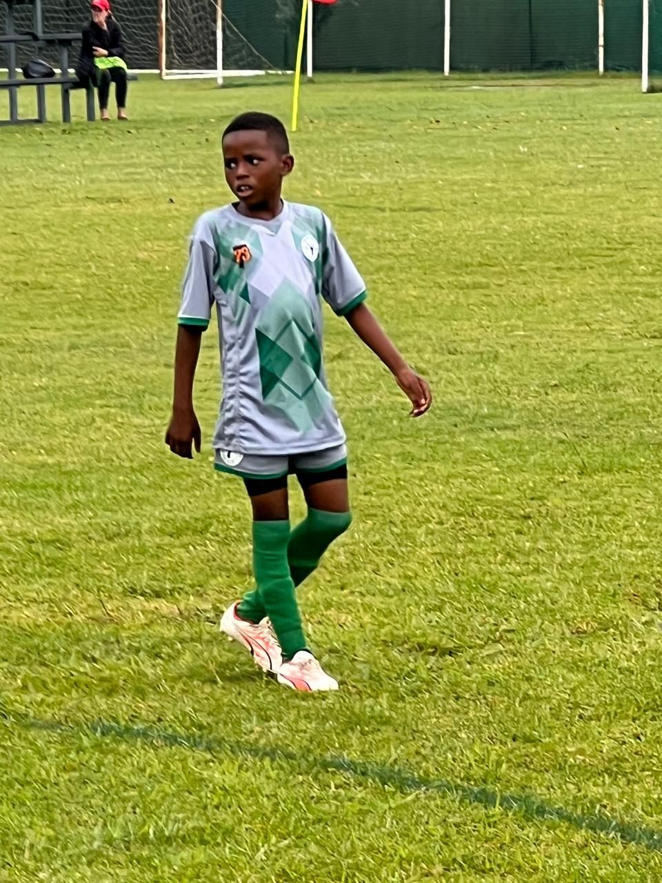 A young soccer player in a grey, patterned jersey and green shorts stands on a grass field, looking to the side.