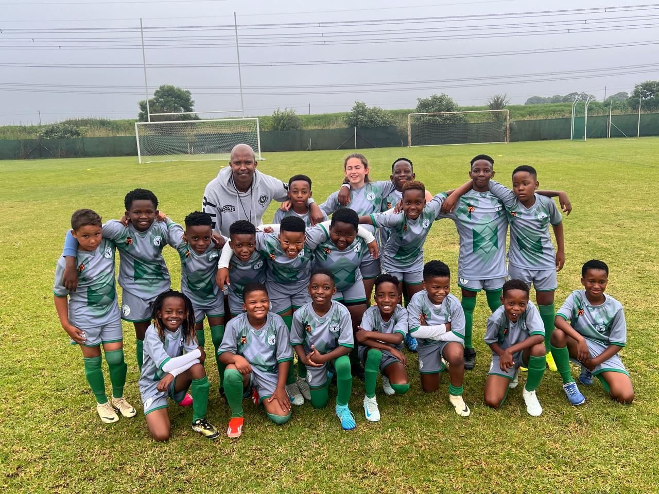 A group of children and a coach in matching green and grey soccer uniforms pose on a grass field.
