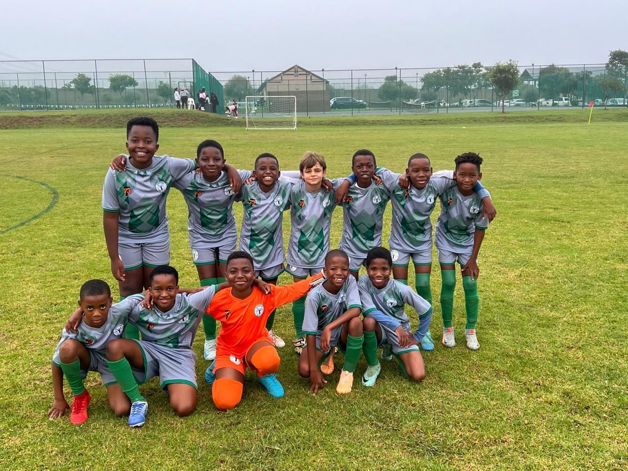 A youth soccer team in green and white uniforms poses for a group photo on a grass field, with one goalie in bright orange.