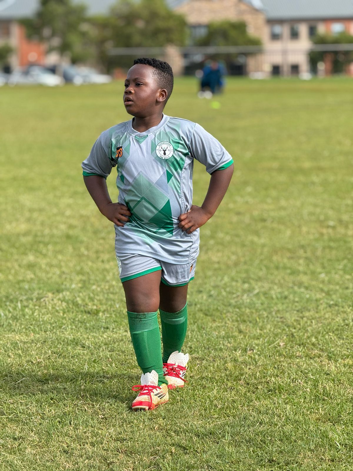 A person in a gray and green soccer uniform stands on a grass field with hands on their hips, looking to the side.