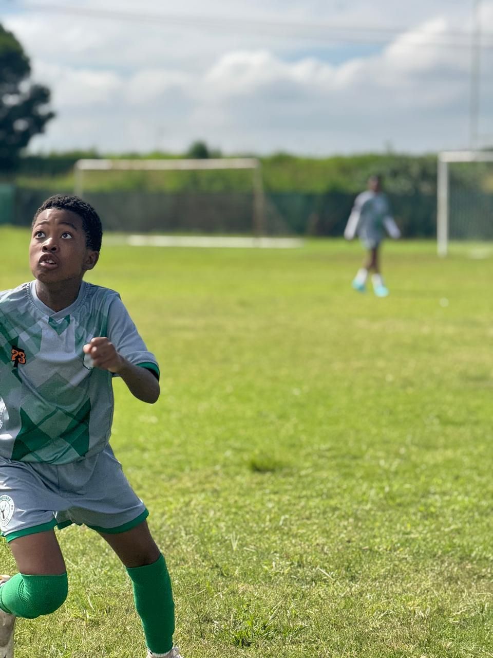 A youth soccer player in a grey and green uniform stands on a grass field, looking upward during a game.