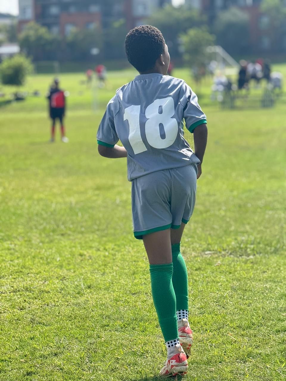 A child in a grey soccer jersey with the number 18 and green socks running on a grass field during a game.